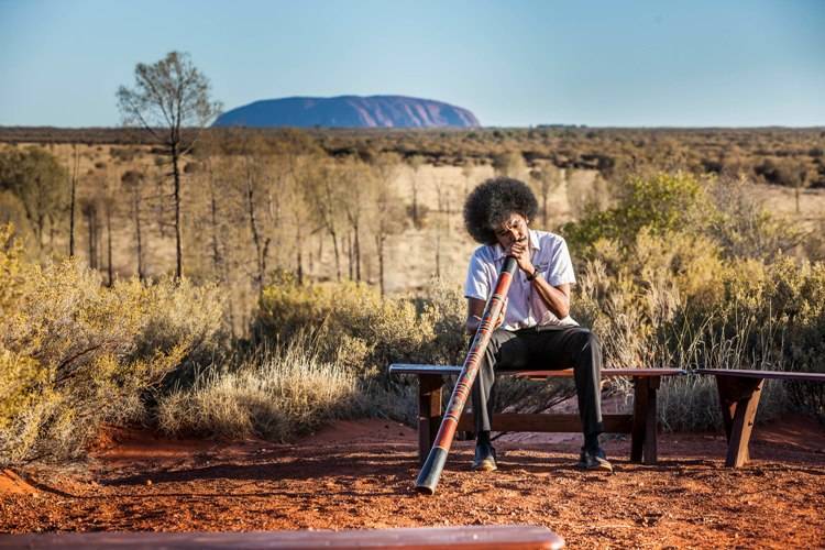 An indigenous Australian playing the wind instrument didgeridoo. In the backdrop is the Ayers Rock